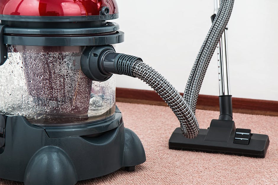 Close-up of a commercial vacuum cleaner with a transparent dust container filled with debris, positioned on a pink carpeted floor in a room with white walls. The vacuum has a black base and a flexible, patterned hose connected to a flat, black cleaning head. The scene illustrates surface cleaning and deep cleaning processes using modern equipment, highlighting the maintenance of hygiene and cleanliness in a domestic or commercial setting, with visible foam and water droplets inside the dust container. The environment appears well-lit, emphasizing the cleanliness of the carpet. This image is associated with Carpet Cleaners Bromley and their professional cleaning services, including emergency carpet cleaning at Bromley North station as indicated by the page title.