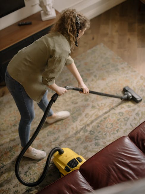 A professional cleaning specialist dressed in protective white coveralls, goggles, and a face mask, is performing surface cleaning with a steam cleaning machine on a beige carpet in a modern living room. The room features a wooden bookshelf with decorative items and books, a tall potted plant, and a white-framed mirror against a white wall. The bright, natural lighting highlights the cleanliness and freshness of the deep-cleaned carpet, which appears fluffy and free of stains or debris. The scene showcases thorough hygiene and routine maintenance associated with domestic cleaning services offered by Carpet Cleaners Bromley, emphasizing effective sanitisation of residential floor surfaces.