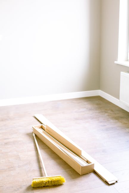 A bright, minimally furnished room with light-colored wooden flooring and white walls. Near the corner by a window that allows natural light to illuminate the space, there is a long, white baseboard a